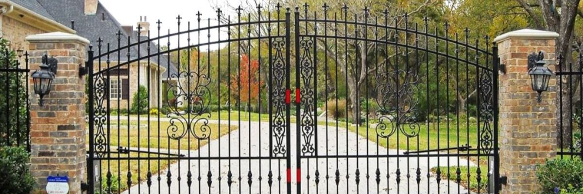 Residential entrance featuring decorative iron gate, brick columns, and landscaped yard with fall foliage
